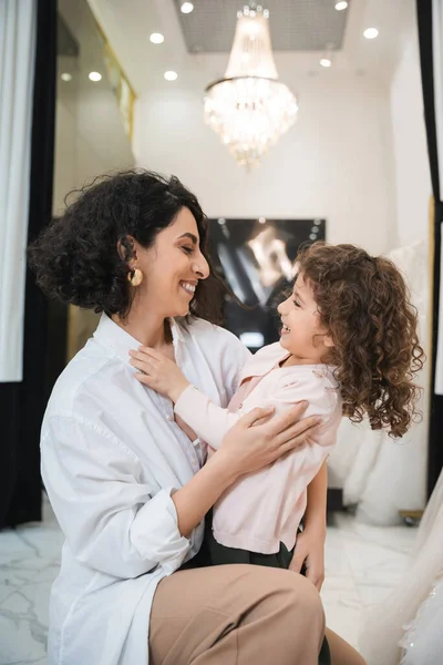 Cheerful middle eastern bride with brunette hair in white shirt hugging happy little girl near white wedding dresses in bridal salon, mother and daughter, bonding, special moment, shopping — Stock Photo