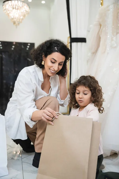 Cheerful middle eastern woman with brunette hair in white shirt and surprised little girl looking inside of shopping bag near white wedding dresses in bridal salon, mother and daughter — Stock Photo