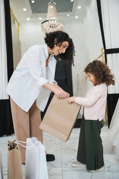 Excited middle eastern woman with brunette hair in white shirt and happy little girl looking inside of shopping bag near wedding dresses in bridal salon, mother and daughter, bridal shopping — Stock Photo