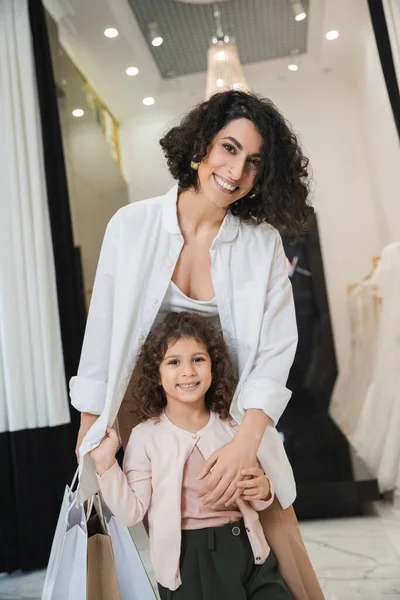Joyful middle eastern woman with brunette hair holding shopping bags and hugging cute little girl while standing near wedding dresses in bridal salon, mother and daughter, bridal shopping — Stock Photo