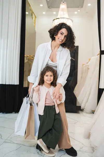 Joyful middle eastern woman with brunette hair holding shopping bags and hugging cute little girl while standing near white wedding dresses in bridal salon, mother and daughter, bridal shopping — Stock Photo