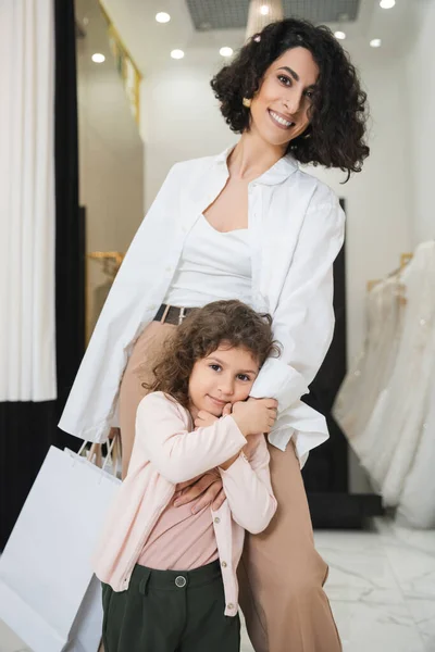 Cute little girl hugging hand of happy middle eastern woman with brunette hair holding shopping bags while standing near wedding dresses in bridal salon, mother and daughter, bridal shopping — Stock Photo