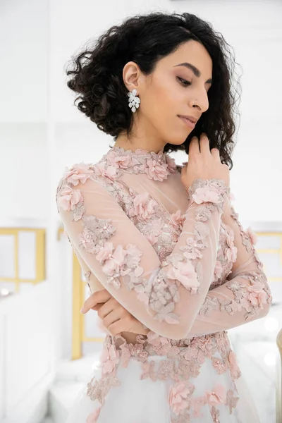 Belle mariée du Moyen-Orient avec des cheveux bruns et ondulés debout dans une magnifique robe de mariée florale et regardant loin à l'intérieur de la boutique de luxe, accents dorés, portrait nuptial — Photo de stock