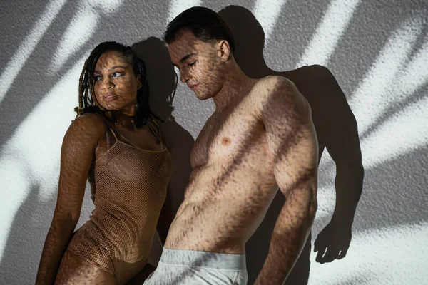 Afrikanisch-amerikanische Frau mit Dreadlocks, im Netzbody neben hemdlosem muskulösen Mann in Unterhosen stehend, junges sexy Paar posiert auf weißem und strukturiertem Hintergrund mit grauen Schatten — Stockfoto