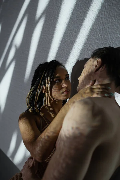 Femme afro-américaine passionnée et sexy avec dreadlocks élégants embrassant jeune homme torse nu près de fond blanc et texturé avec des ombres grises, moment intime de couple multiethnique — Photo de stock