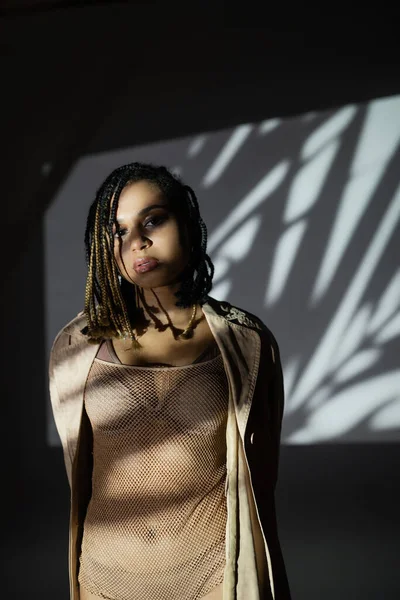 Charming and sexy african american woman with stylish dreadlocks, in lingerie, net bodysuit and jacket looking at camera while posing in light on black and white background with white shadows — Stock Photo
