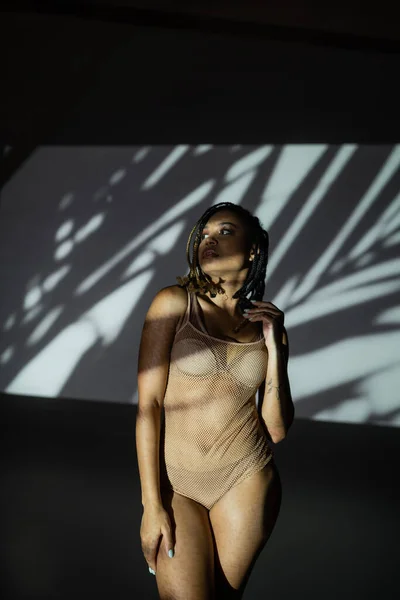 Молода і гламурна афроамериканська жінка в сексуальній білизні і сітчастому боді стоїть і дивиться в сторону, стоячи у світлі на абстрактному чорно-білому тлі з сірими тінями — стокове фото