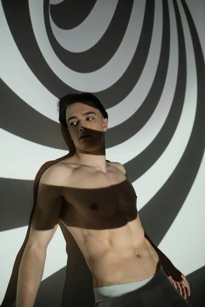 Sexy, jovem e bonito homem com tronco muscular e corpo sem camisa olhando para longe enquanto em pé e posando em cuecas em abstrato fundo preto e branco com projeção em espiral — Fotografia de Stock