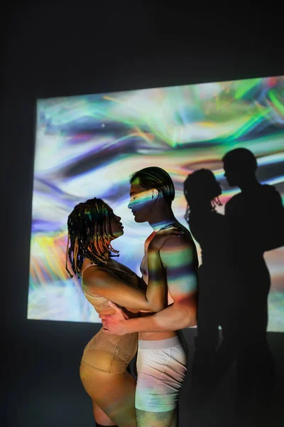 Side view of young, shirtless and muscular man embracing appealing african american woman with dreadlocks, in net bodysuit on black background with colorful projection and lighting effects — Stock Photo