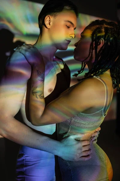 Muskulöser, hemdloser Mann umarmt charmante Afroamerikanerin mit stylischen Dreadlocks, im Netzbody auf Hintergrund mit farbenfrohen Projektionen und Lichteffekten — Stockfoto