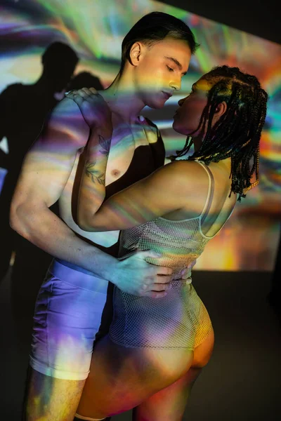 Momento íntimo de homem sem camisa e musculoso abraçando mulher americana africana tatuada com dreadlocks, em bodysuit líquido em fundo preto com projeção colorida e efeitos de iluminação — Fotografia de Stock