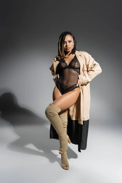Full length of expressive african american woman with dreadlocks, in black lace bodysuit, beige trench coat and over knee boots posing with hands on hips and looking at camera on grey background — Stock Photo