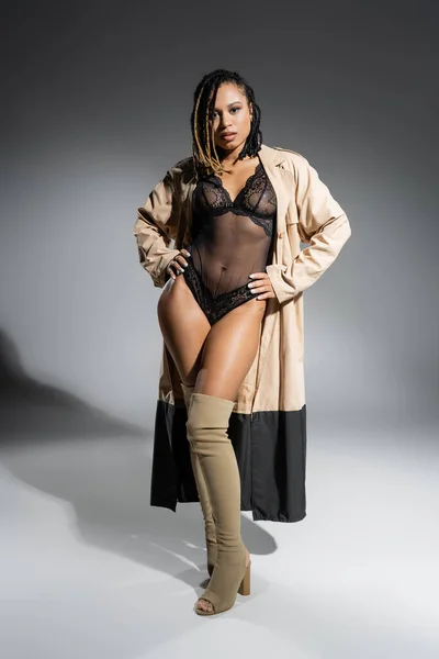 Full length of african american woman with dreadlocks, in trendy beige coat, sexy black lace bodysuit and over knee boots standing with hands on hips and looking at camera on grey background — Stock Photo
