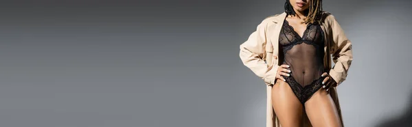 Vista parziale della graziosa e sexy donna afroamericana in trench beige alla moda e lingerie nera in pizzo in posa con le mani sui fianchi su sfondo grigio, banner — Foto stock