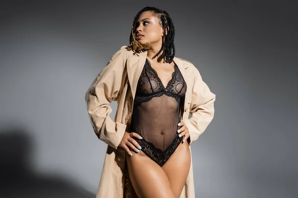 Expresiva mujer afroamericana con rastas, en sexy traje de encaje negro y elegante gabardina beige posando con las manos en las caderas y mirando hacia otro lado sobre fondo gris - foto de stock