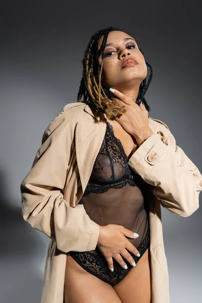 Provocativa mujer afroamericana con rastas, en traje de encaje negro y gabardina beige de moda tocando el cuello mientras está de pie en pose provocativa y mirando a la cámara sobre fondo gris - foto de stock