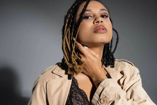 Sassy and charming african american woman with smokey makeup and dreadlocks posing in black lace lingerie and beige trench coat while standing in provocative pose on grey background — Stock Photo