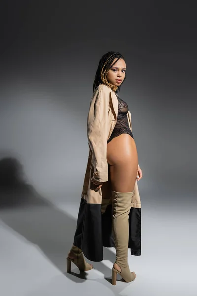Full length of seductive and trendy african american woman in black lace bodysuit, beige trench coat and over knee boots standing and looking at camera on grey background — Stock Photo