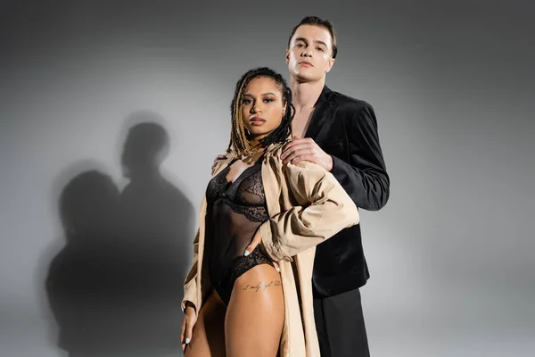 Fashionable interracial couple, young man in black silk blazer, african american woman in lace bodysuit and beige trench coat looking at camera on grey background with shadows — Stock Photo