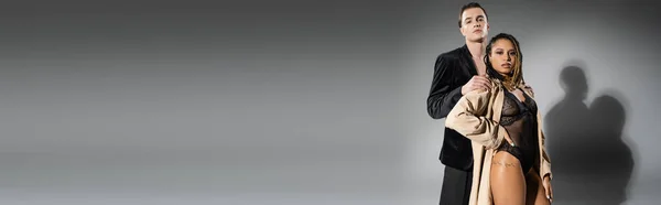 Сексуальна афроамериканська жінка в мереживному костюмі і бежева траншея пальто позує з рукою на стегні біля красивого чоловіка в чорному шовковому блістері на сірому фоні, банер — стокове фото