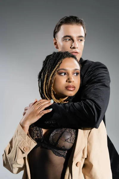 Young good looking man in black silk blazer embracing charming african american woman with dreadlocks wearing lace lingerie and beige trench coat while looking away on grey background — Stock Photo