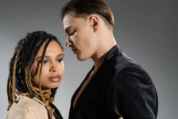 Young and handsome man in black stylish blazer standing near charming african american woman with dreadlocks and smokey makeup looking away on grey background — Stock Photo