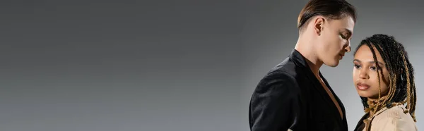 Jovem e cativante mulher americana africana com dreadlocks elegantes olhando para longe perto homem na moda em blazer de seda preta no fundo cinza, banner — Fotografia de Stock