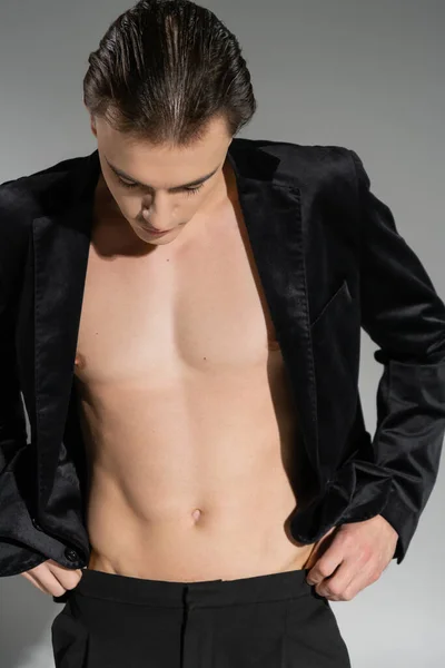 Young and good looking man with brunette hair, wearing black silk blazer on shirtless muscular body, adjusting pants while standing on grey background — Stock Photo
