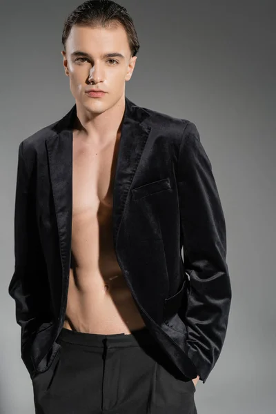 Homem auto-confiante e atraente vestindo blazer de seda preta no corpo muscular sem camisa, de pé com as mãos nos bolsos e olhando para a câmera no fundo cinza — Fotografia de Stock