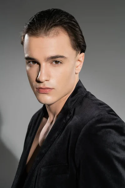 Portrait of young and stylish man with confident face expression and brunette hair looking at camera while posing in black silk blazer on grey background — Stock Photo