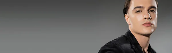 Porträt eines selbstbewussten und jungen selbstbewussten Mannes mit brünetten Haaren, der in die Kamera blickt, während er im schwarzen Seidenblazer auf grauem Hintergrund mit Kopierraum posiert, Banner — Stockfoto