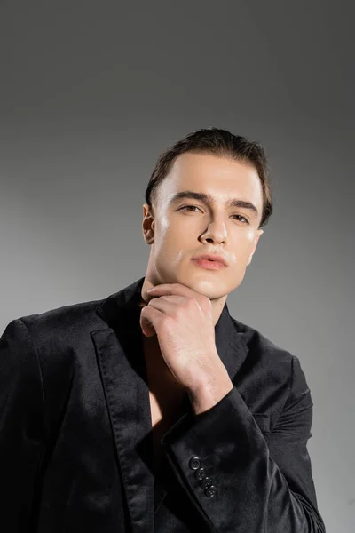 Portrait of young, self-assured and fashionable man wearing black silk blazer and looking at camera while posing with hand near chin on grey background — Stock Photo