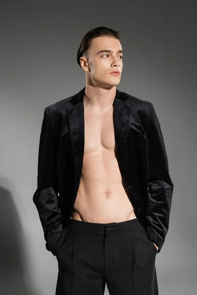 Junger, sexy und modischer Mann mit schwarzem Seidenblazer am schulterfreien muskulösen Oberkörper, der wegschaut, während er mit den Händen in Hosentaschen auf grauem Hintergrund posiert — Stockfoto