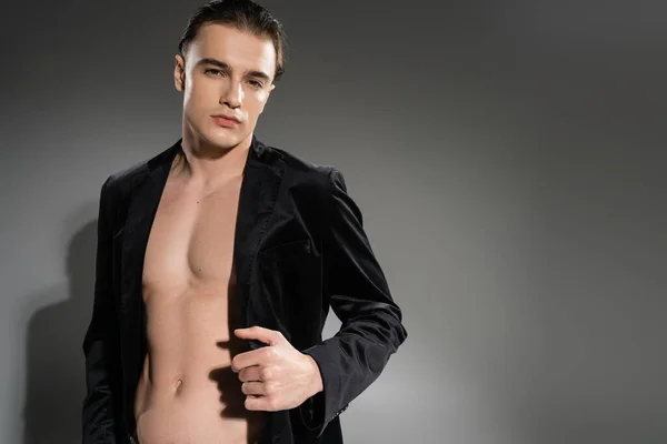 Young, confident and good looking man with brunette hair, wearing black and silk blazer on shirtless muscular body while looking at camera on grey background — Stock Photo