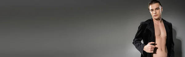 Giovane, bello e alla moda uomo con i capelli castani, indossa blazer di seta nera sul corpo senza maglietta, posa e guardando la fotocamera su sfondo grigio, banner — Foto stock