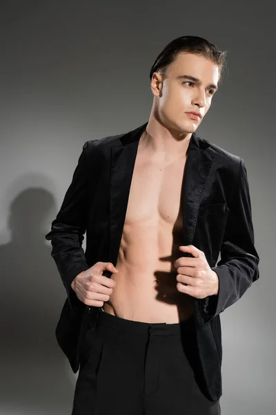 Hombre joven, seguro de sí mismo y de moda con el pelo moreno posando en blazer negro y de seda sobre el cuerpo musculoso sin camisa y mirando hacia otro lado sobre fondo gris - foto de stock