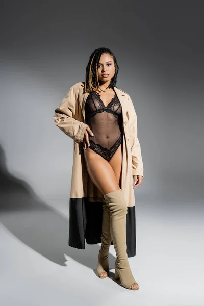 Full length of provocative and attractive african american woman in black lace bodysuit, beige trench coat and over knee boots posing with hand on hip and looking at camera on grey background — Stock Photo