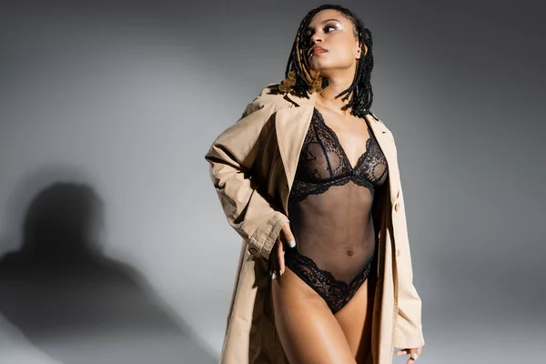 Sexy und selbstbewusste afrikanisch-amerikanische Frau mit Dreadlocks, in modischem beigen Blazer und schwarzem Spitzenbody, stehend und wegschauend auf grauem Hintergrund — Stockfoto