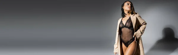 Femme afro-américaine enchanteresse avec dreadlocks élégants, dans un body en dentelle noire debout et regardant loin sur fond gris avec espace de copie, bannière — Photo de stock