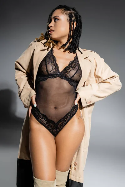 Сексуальная и модная африканская американка с дредами, в черном кружевном костюме и бежевом плаще смотрит в сторону, позируя руками на бедрах на сером фоне — стоковое фото