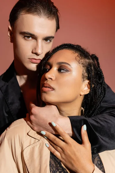 Young charismatic man in black silk blazer looking at camera while hugging neck of sexy african american woman with dreadlocks and smokey makeup on pinkish beige background — Stock Photo