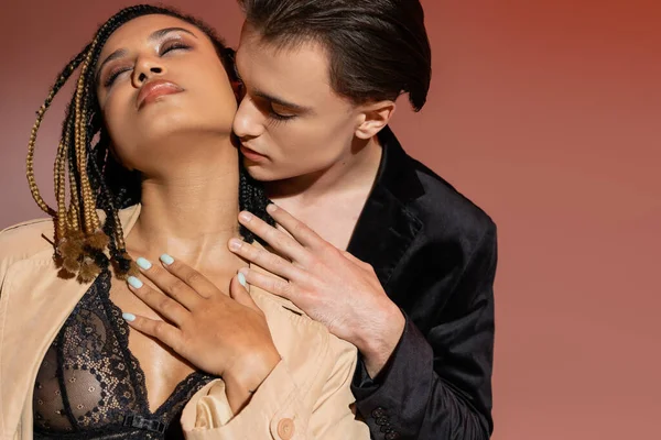 Hombre joven y sexy en chaqueta de seda negra seduciendo y abrazando a mujer afroamericana con rastas, ojos cerrados y maquillaje ahumado, en lencería de encaje y gabardina sobre fondo beige rosado - foto de stock
