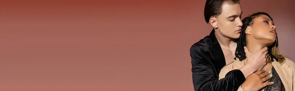 Homme à la mode et beau en veste de soie noire embrassant le cou d'une femme afro-américaine passionnée avec dreadlocks vêtus de lingerie en dentelle et trench coat sur fond beige rosé, bannière — Stock Photo