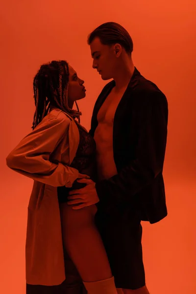 Боковой вид сексуального мужчины в черном блейзере на теле без рубашки, обнимающего провокационную африканскую женщину в кружевном костюме и бежевом траншеи стоимостью на оранжевом фоне с эффектом красного освещения — стоковое фото