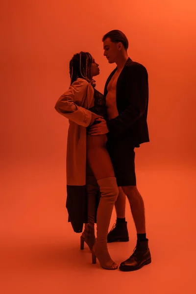 Volle Länge des Mannes in schwarzem Blazer und Shorts verführt sexy afrikanisch-amerikanische Frau in Trenchcoat, Spitzenbody und Overknee-Stiefel stehend mit der Hand an der Hüfte auf orangefarbenem Hintergrund mit roter Beleuchtung — Stockfoto