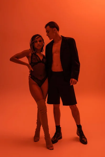 Full length of stylish man in blazer and shorts embracing african american woman in black lace bodysuit and over knee boots standing with hand on waist on orange background with red lighting effect — Stock Photo