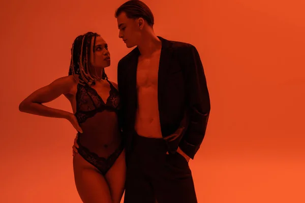 Glamouröser Mann im schwarzen Blazer auf hemdlosem Körper und sexy provokante afrikanisch-amerikanische Frau mit Spitzenbody, die einander auf orangefarbenem Hintergrund mit rotem Lichteffekt anschaut — Stockfoto