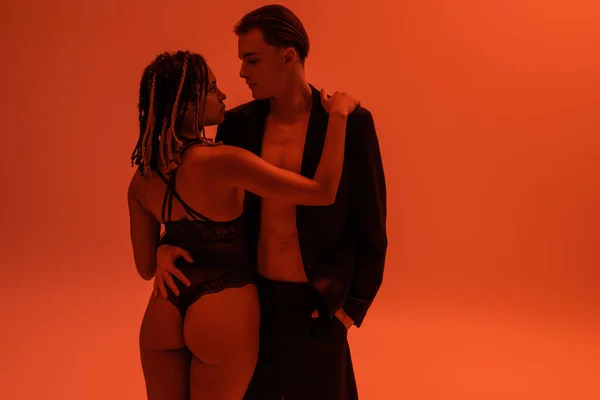 Young and stylish man standing with hand in pocket of black shorts and hugging appealing african american woman with sexy buttocks, wearing lace bodysuit on orange background with red lighting — Stock Photo