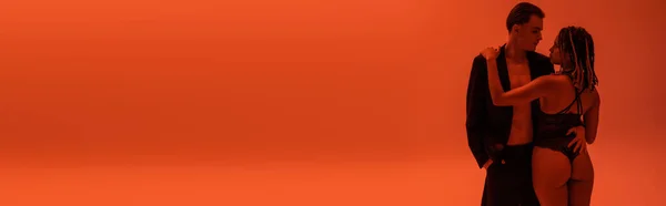Hombre joven y sexy en blazer en cuerpo sin camisa de pie con la mano en el bolsillo y abrazando a la seductora mujer afroamericana en traje de encaje negro y sobre fondo naranja con iluminación roja, pancarta - foto de stock