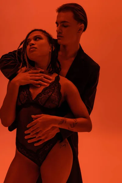 Young and self-assured man in blazer hugging provocative and sexy african american woman with dreadlocks wearing black lace bodysuit on orange background with red lighting effect — Stock Photo